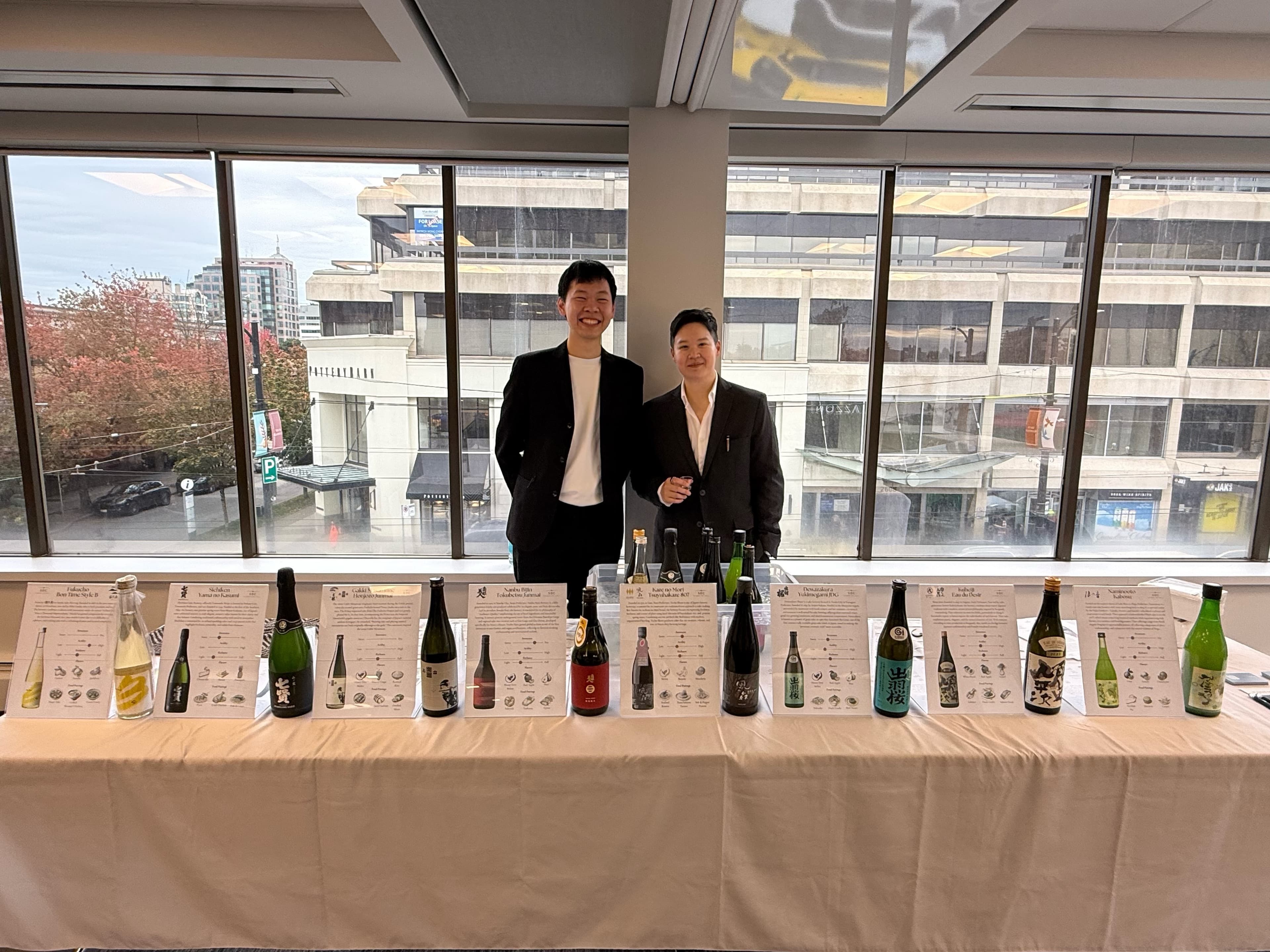 Spencer and Andre behind a long tasting table lined with premium sake bottles and tasting cards