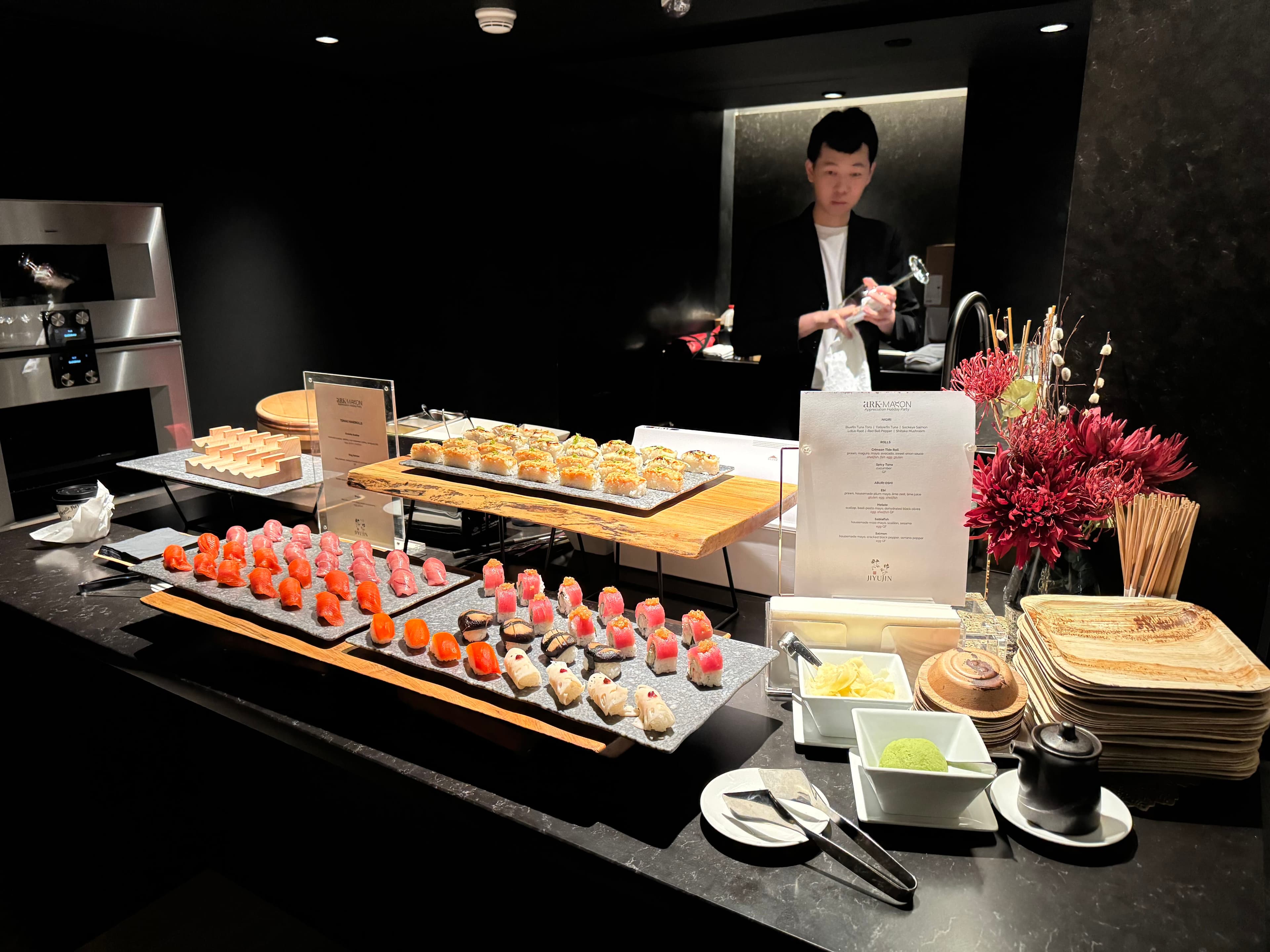 A chef plating nigiri behind a counter of sushi platters, tasting menus, and a red floral arrangement